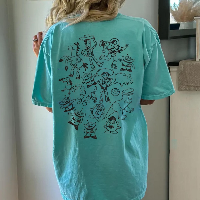 May include: A light blue t-shirt featuring a black graphic of cartoon characters. The design includes a cowboy, astronaut, dinosaur, and other characters. The shirt is made of a soft material.