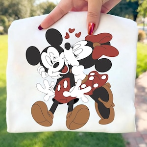 May include: White sweatshirt featuring a graphic of Mickey Mouse and Minnie Mouse embracing and kissing, with hearts above them. Mickey is wearing red shorts and brown shoes. Minnie is wearing a red polka dot skirt and bow. The design is centered on the sweatshirt.