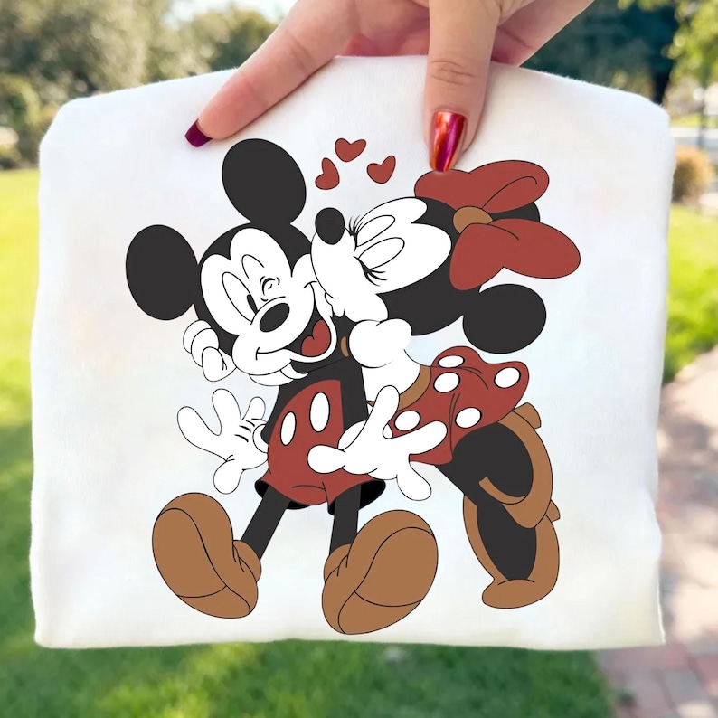 May include: White sweatshirt featuring a graphic of Mickey Mouse and Minnie Mouse embracing and kissing, with hearts above them. Mickey is wearing red shorts and brown shoes. Minnie is wearing a red polka dot skirt and bow. The design is centered on the sweatshirt.