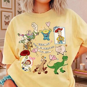 May include: A pale yellow t-shirt featuring a graphic print of Toy Story characters including Buzz Lightyear, Woody, Jessie, Rex, Slinky Dog, Hamm, and an Alien. The text "YOU'VE GOT A FRIEND IN ME" is arranged in a circle in the center of the design.