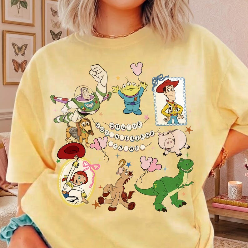 May include: A pale yellow t-shirt featuring a graphic print of Toy Story characters including Buzz Lightyear, Woody, Jessie, Rex, Slinky Dog, Hamm, and an Alien. The text "YOU'VE GOT A FRIEND IN ME" is arranged in a circle in the center of the design.