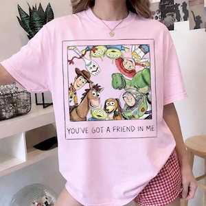 K&ouml;nnte beinhalten: Hellrosa T-Shirt mit einem gerahmten Aufdruck von Toy Story-Charakteren, darunter Woody und Buzz Lightyear. Der Text "YOU'VE GOT A FRIEND IN ME" ist unter dem Bild gedruckt.