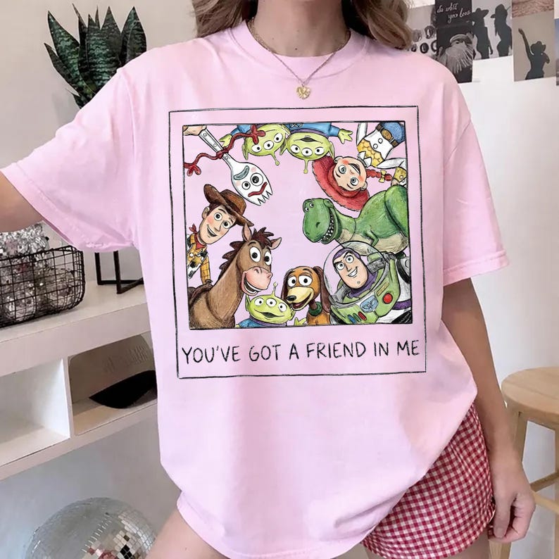 K&ouml;nnte beinhalten: Hellrosa T-Shirt mit einem gerahmten Aufdruck von Toy Story-Charakteren, darunter Woody und Buzz Lightyear. Der Text "YOU'VE GOT A FRIEND IN ME" ist unter dem Bild gedruckt.