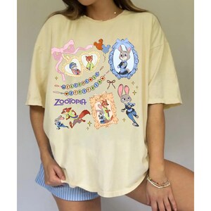 May include: A light yellow t-shirt featuring various cartoon images and the word "Zootopia". The shirt has a crew neck and short sleeves. The graphics include framed character portraits and decorative elements.