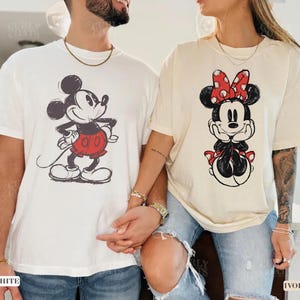 May include: Two t-shirts featuring Disney characters. One is white with a Mickey Mouse graphic in black, red, and white. The other is ivory with a Minnie Mouse graphic in black, red, and white. Both shirts have short sleeves.