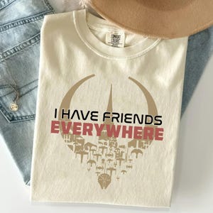 以下が含まれることがあります： クリーム色の半袖Tシャツ。円形に配置された様々な宇宙船のベージュ色のグラフィックが描かれています。宇宙船のグラフィックの中央には、黒い文字で「I HAVE FRIENDS」、赤い文字で「EVERYWHERE」とプリントされています。