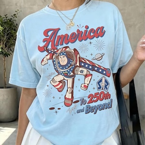 May include: A light blue t-shirt featuring a graphic of a space ranger character in a red, white, and blue suit. The text reads America To 250th and Beyond, accompanied by illustrations of fireworks, a planet, and a star-shaped logo with a rocket.