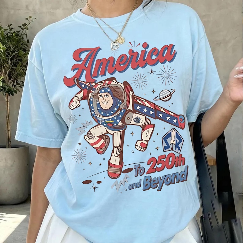 May include: A light blue t-shirt featuring a graphic of a space ranger character in a red, white, and blue suit. The text reads America To 250th and Beyond, accompanied by illustrations of fireworks, a planet, and a star-shaped logo with a rocket.