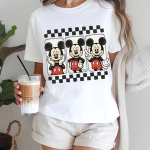 May include: White t-shirt featuring a graphic of Mickey Mouse in three different poses. The design includes a black and white checkered border. Mickey is wearing his signature red shorts and black gloves.