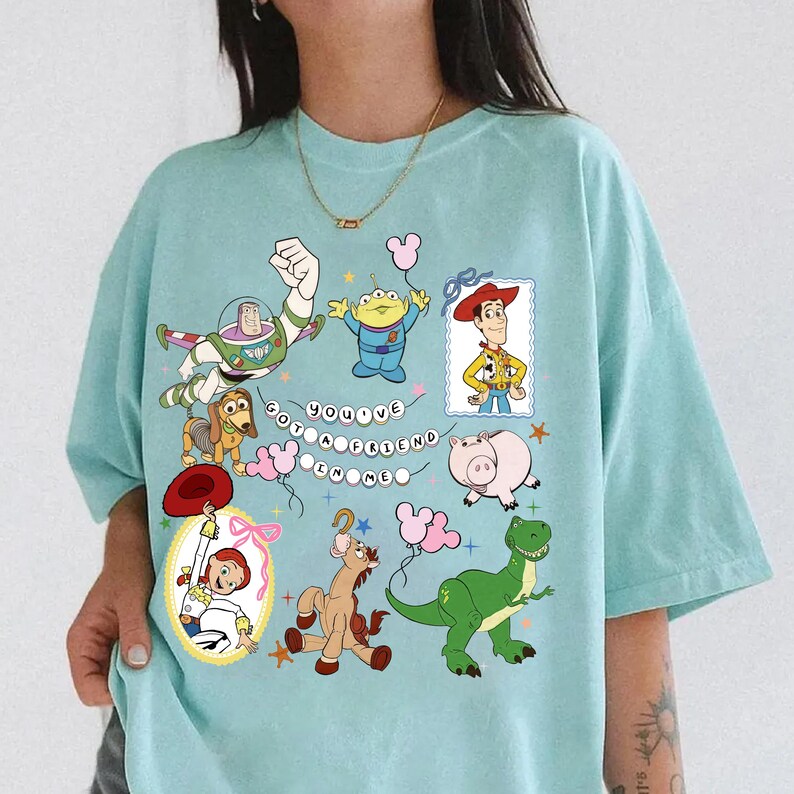 May include: A light blue t-shirt featuring a graphic print of various animated characters including a space ranger, a cowboy, a cowgirl, a dinosaur, a pig, a dog, and an alien. The text "YOU'VE GOT A FRIEND IN ME" is printed in the center with small stars.
