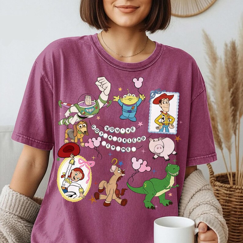 May include: A plum-colored short-sleeve t-shirt featuring a graphic print of Toy Story characters including Buzz Lightyear, Woody, Jessie, Rex, Slinky Dog, Hamm, and an alien. The text "YOU'VE GOT A FRIEND IN ME" is displayed in the center.