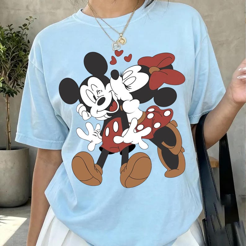 May include: Light blue t-shirt featuring a graphic of Mickey and Minnie Mouse embracing, with hearts floating above them. Mickey wears red shorts and brown shoes, while Minnie has a red bow and polka-dot skirt. The shirt is a casual, unisex style.
