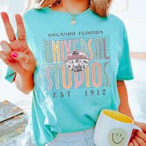 May include: A turquoise t-shirt with the text "ORLANDO FLORIDA UNIVERSAL STUDIOS EST. 1912" in colorful block letters. The shirt features a graphic of the Universal Studios logo. A white mug with a yellow interior and a smiley face is also visible.