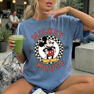May include: A pale blue t-shirt with a Mickey Mouse design. The graphic features the words "Mickey Mouse" in red, with Mickey Mouse in the center. The shirt also has the text "EST. 1928".