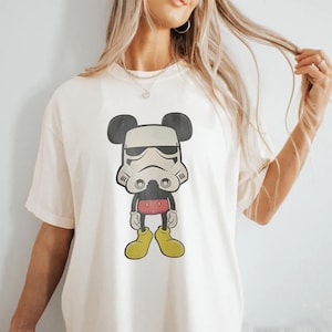 May include: A cream-coloured t-shirt with a cartoon character mashup design. The graphic combines Mickey Mouse's silhouette with a Stormtrooper helmet. The character wears red shorts, yellow shoes, and black gloves. A fun, pop culture-themed graphic tee.