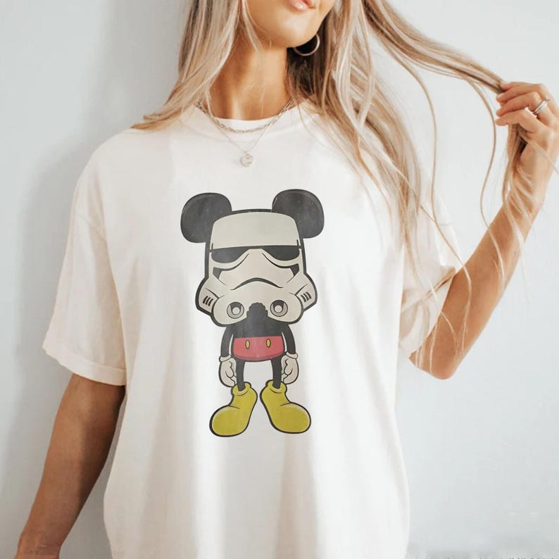 May include: A cream-coloured t-shirt with a cartoon character mashup design. The graphic combines Mickey Mouse's silhouette with a Stormtrooper helmet. The character wears red shorts, yellow shoes, and black gloves. A fun, pop culture-themed graphic tee.