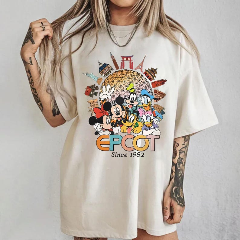 Pu&ograve; includere: T-shirt color crema con una grafica colorata di personaggi Disney, tra cui Topolino e Minnie, Paperino e Pippo, con la scritta "EPCOT Since 1982". La grafica include monumenti iconici.