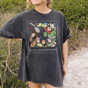 K&ouml;nnte beinhalten: Dunkelgraues T-Shirt mit einer farbenfrohen Grafik von Figuren aus dem Film Toy Story. Die Grafik ist in einem wei&szlig;en Rechteck gerahmt und enth&auml;lt den Text "YOU'VE GOT A FRIEND IN ME". Das Shirt hat kurze &Auml;rmel und eine lockere Passform.