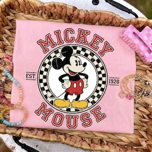 May include: Pale pink t-shirt with a Mickey Mouse graphic in a circular design, framed by a black and white chequered border. The words "MICKEY MOUSE" are arched above and below the character, with "EST. 1928" on either side. Bracelets and a pink hair clip are also visible.