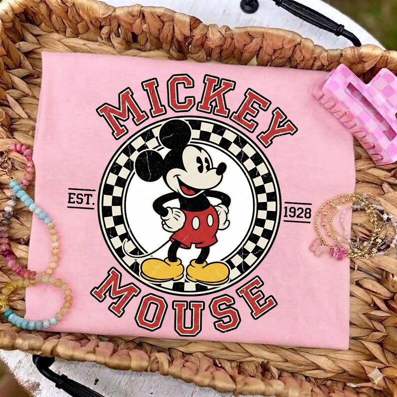 May include: Pale pink t-shirt with a Mickey Mouse graphic in a circular design, framed by a black and white chequered border. The words "MICKEY MOUSE" are arched above and below the character, with "EST. 1928" on either side. Bracelets and a pink hair clip are also visible.