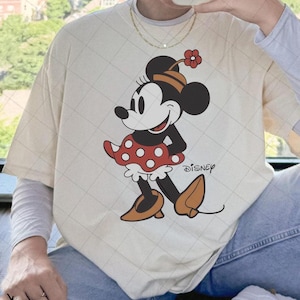 May include: Cream-colored t-shirt featuring a graphic of Minnie Mouse in a red polka-dot dress, brown shoes, and a flower on her head. The word "DISNEY" is printed on the shirt. The shirt is worn by a person.