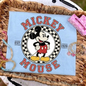 May include: Pale blue t-shirt with a Mickey Mouse graphic. The design features the words "Mickey Mouse" in red, a black and white chequered circle, and the year "1928". The shirt is surrounded by bracelets and a hair clip.
