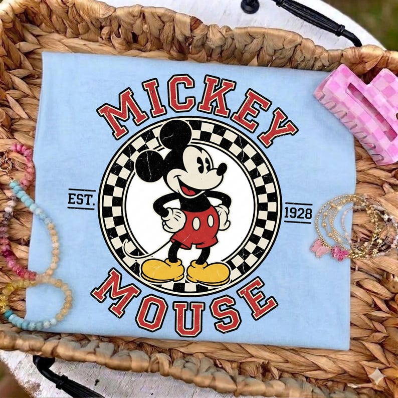 May include: Pale blue t-shirt with a Mickey Mouse graphic. The design features the words "Mickey Mouse" in red, a black and white chequered circle, and the year "1928". The shirt is surrounded by bracelets and a hair clip.