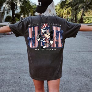 May include: Dark grey t-shirt with a patriotic "USA" design in red and white stripes and blue stars. The design includes a cartoon character in patriotic clothing, with the text "250th Anniversary" and "1776 2026".
