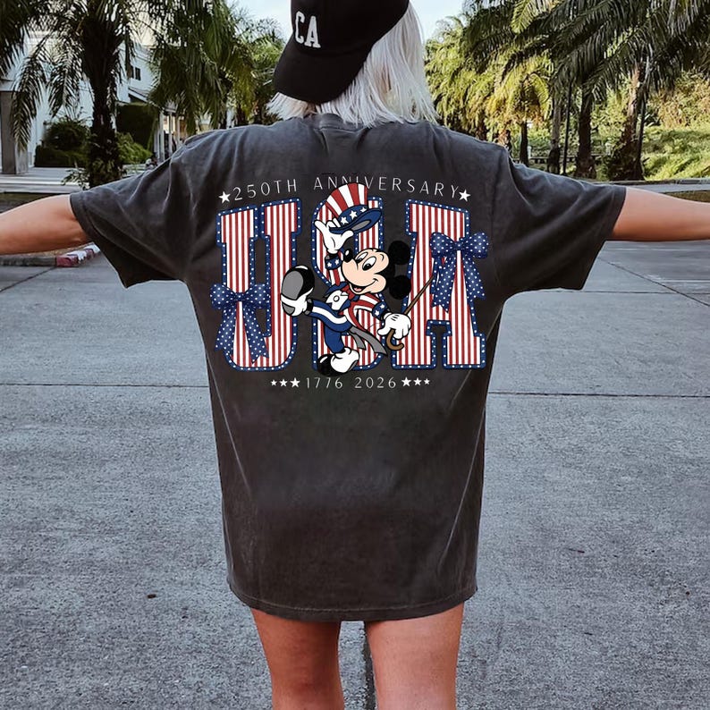 May include: Dark grey t-shirt with a patriotic "USA" design in red and white stripes and blue stars. The design includes a cartoon character in patriotic clothing, with the text "250th Anniversary" and "1776 2026".