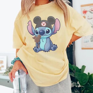May include: A light yellow t-shirt featuring a cartoon illustration of Stitch, wearing a Mickey Mouse hat and holding a chocolate ice cream bar. The graphic is in shades of blue, black, and brown. The shirt is a casual style.