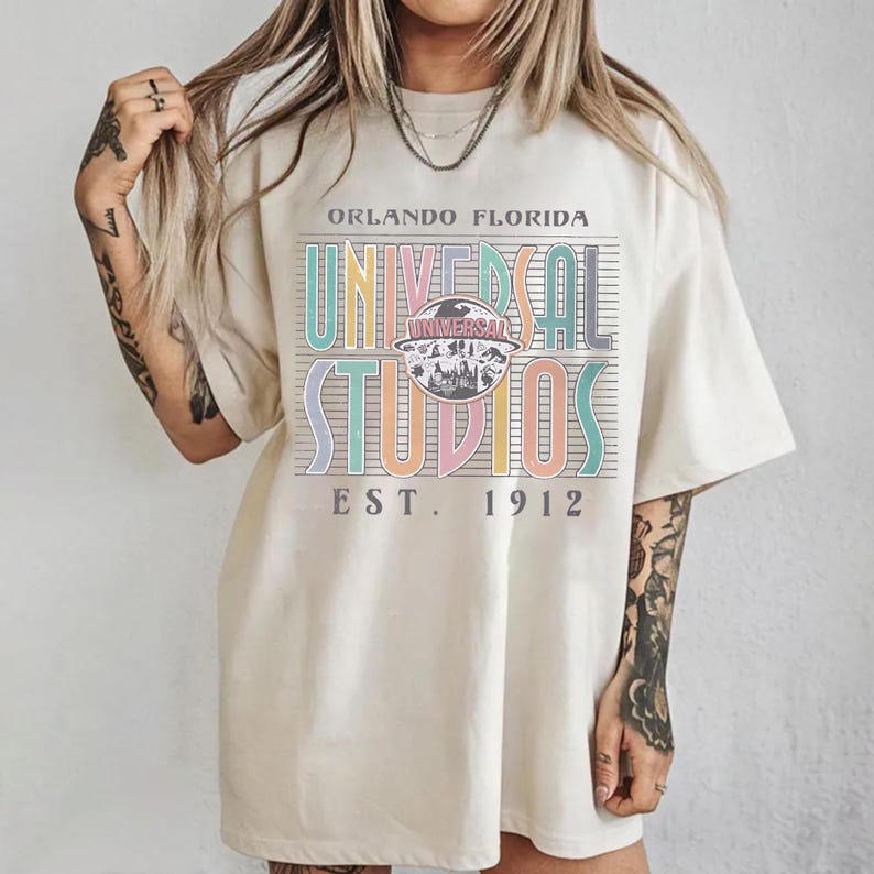 May include: Cream-colored t-shirt featuring the text "ORLANDO FLORIDA UNIVERSAL STUDIOS EST. 1912" in a colorful, retro design. The graphic includes a central emblem with a silhouette of a cityscape.
