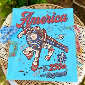 May include: A light blue t-shirt featuring a graphic of Buzz Lightyear in a red, white, and blue space suit. The text reads America To 250th and Beyond, accompanied by starbursts, a planet, and a space ranger logo in red, white, and blue colors.