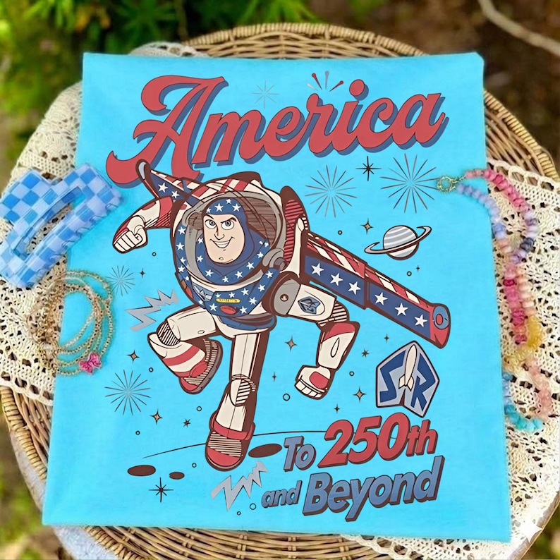 May include: A light blue t-shirt featuring a graphic of Buzz Lightyear in a red, white, and blue space suit. The text reads America To 250th and Beyond, accompanied by starbursts, a planet, and a space ranger logo in red, white, and blue colors.
