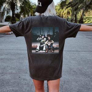 Puede incluir: Camiseta gris oscuro con un estampado rectangular en la espalda. El estampado representa a dos personajes de dibujos animados, uno con un traje espacial y otro con un atuendo de vaquero, sentados juntos.