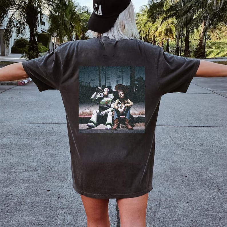 Puede incluir: Camiseta gris oscuro con un estampado rectangular en la espalda. El estampado representa a dos personajes de dibujos animados, uno con un traje espacial y otro con un atuendo de vaquero, sentados juntos.