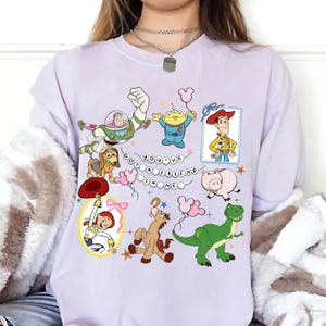 May include: A light purple t-shirt featuring a graphic print of Toy Story characters including Buzz Lightyear, Woody, Jessie, Rex, Slinky Dog, Hamm, Bullseye, and an Alien. The text "YOU'VE GOT A FRIEND IN ME" is displayed in the center with small stars.