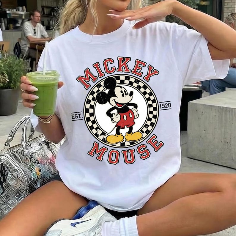 May include: White t-shirt with a Mickey Mouse graphic. The design features a circular emblem with Mickey, the words "Mickey Mouse" in red, and the year "1928". The shirt has a crew neck and short sleeves.