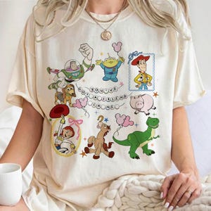 May include: A cream-colored short-sleeve t-shirt featuring a colorful graphic print of various animated characters including a space ranger, a cowboy, a cowgirl, a green dinosaur, and a pig. The text "YOU'VE GOT A FRIEND IN ME" is centered.