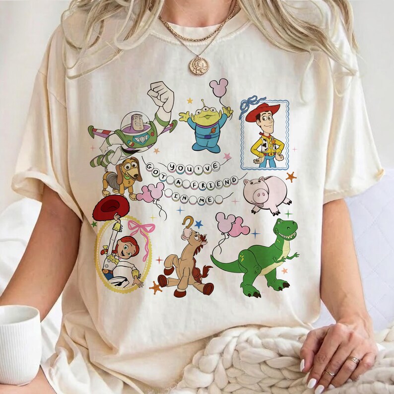 May include: A cream-colored short-sleeve t-shirt featuring a colorful graphic print of various animated characters including a space ranger, a cowboy, a cowgirl, a green dinosaur, and a pig. The text "YOU'VE GOT A FRIEND IN ME" is centered.