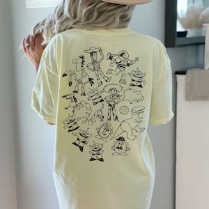 May include: A light yellow t-shirt featuring a black graphic of cartoon characters. The design includes a cowboy, astronaut, and other characters. The shirt is a relaxed fit.