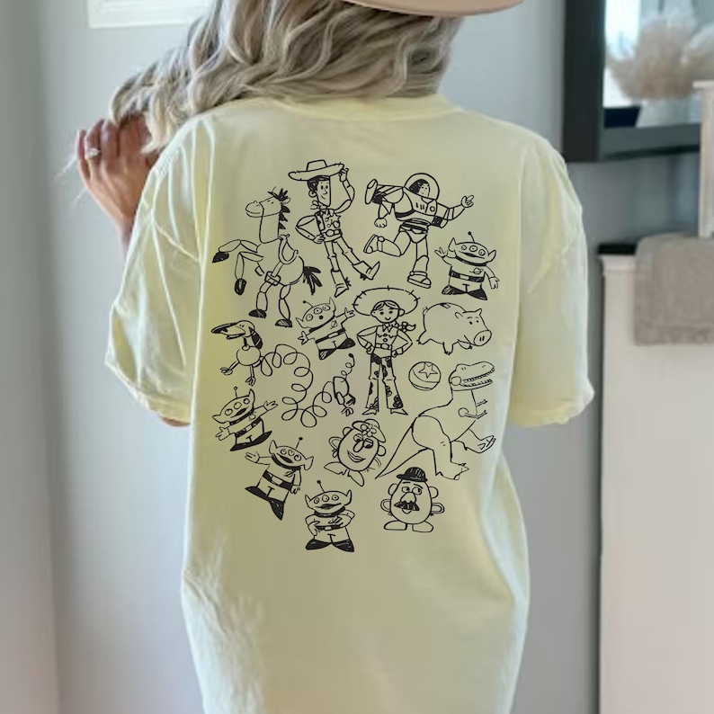 May include: A light yellow t-shirt featuring a black graphic of cartoon characters. The design includes a cowboy, astronaut, and other characters. The shirt is a relaxed fit.