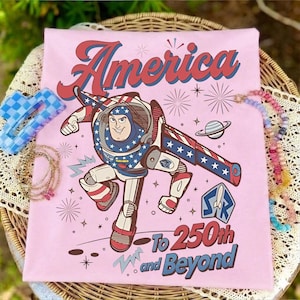 May include: A light pink t-shirt featuring a graphic of a space ranger character in a red, white, and blue suit. The text reads America To 250th and Beyond, accompanied by illustrations of fireworks, a planet, and a space ranger logo emblem.
