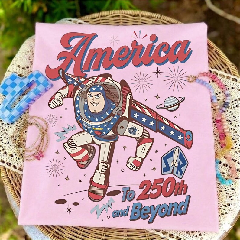 May include: A light pink t-shirt featuring a graphic of a space ranger character in a red, white, and blue suit. The text reads America To 250th and Beyond, accompanied by illustrations of fireworks, a planet, and a space ranger logo emblem.