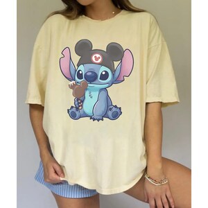 May include: A light yellow t-shirt featuring a cartoon illustration of Stitch from Lilo & Stitch. Stitch is blue with large ears, wearing a Mickey Mouse hat and holding a brown ice cream treat. The shirt has short sleeves.