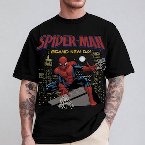 May include: A black short-sleeve t-shirt featuring a graphic print of Spider-Man in a crouching pose. The text reads SPIDER-MAN BRAND NEW DAY above the character, with a city skyline background, a white moon, and a web design in the foreground.