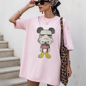 May include: A light pink oversized t-shirt with a cartoon mashup of Mickey Mouse and a Stormtrooper. The graphic is in black, white, red, and yellow. The shirt has short sleeves.