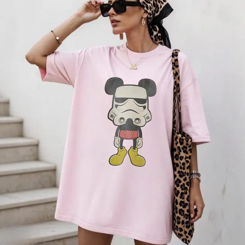 May include: A light pink oversized t-shirt with a cartoon mashup of Mickey Mouse and a Stormtrooper. The graphic is in black, white, red, and yellow. The shirt has short sleeves.