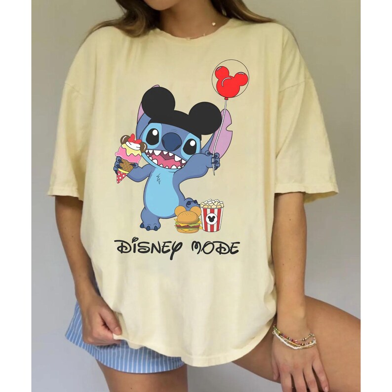 May include: A light yellow t-shirt featuring a cartoon character holding an ice cream cone, a balloon, and other snacks. The text "DISNEY MODE" is printed in black script below the character.