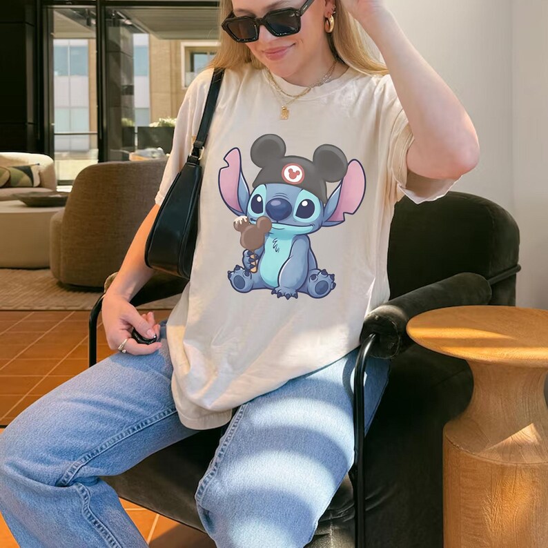 May include: Cream-colored t-shirt featuring a cartoon illustration of Stitch wearing a Mickey Mouse hat and holding a brown ice cream cone. The graphic is centered on the shirt. The shirt is a relaxed fit.