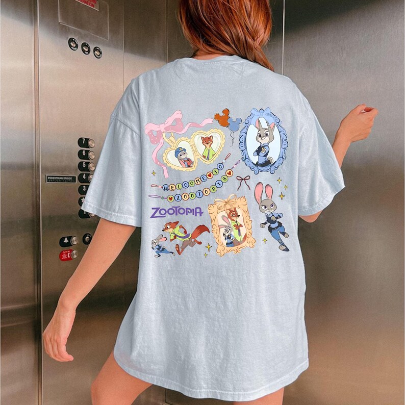 May include: Light blue t-shirt featuring colorful illustrations of characters from the animated film "Zootopia." The design includes framed portraits, a banner with the film's title, and various character graphics. The shirt has short sleeves.